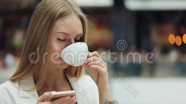 迷人的年轻女子一手拿着智能手机，一手拿着咖啡坐在咖啡馆里